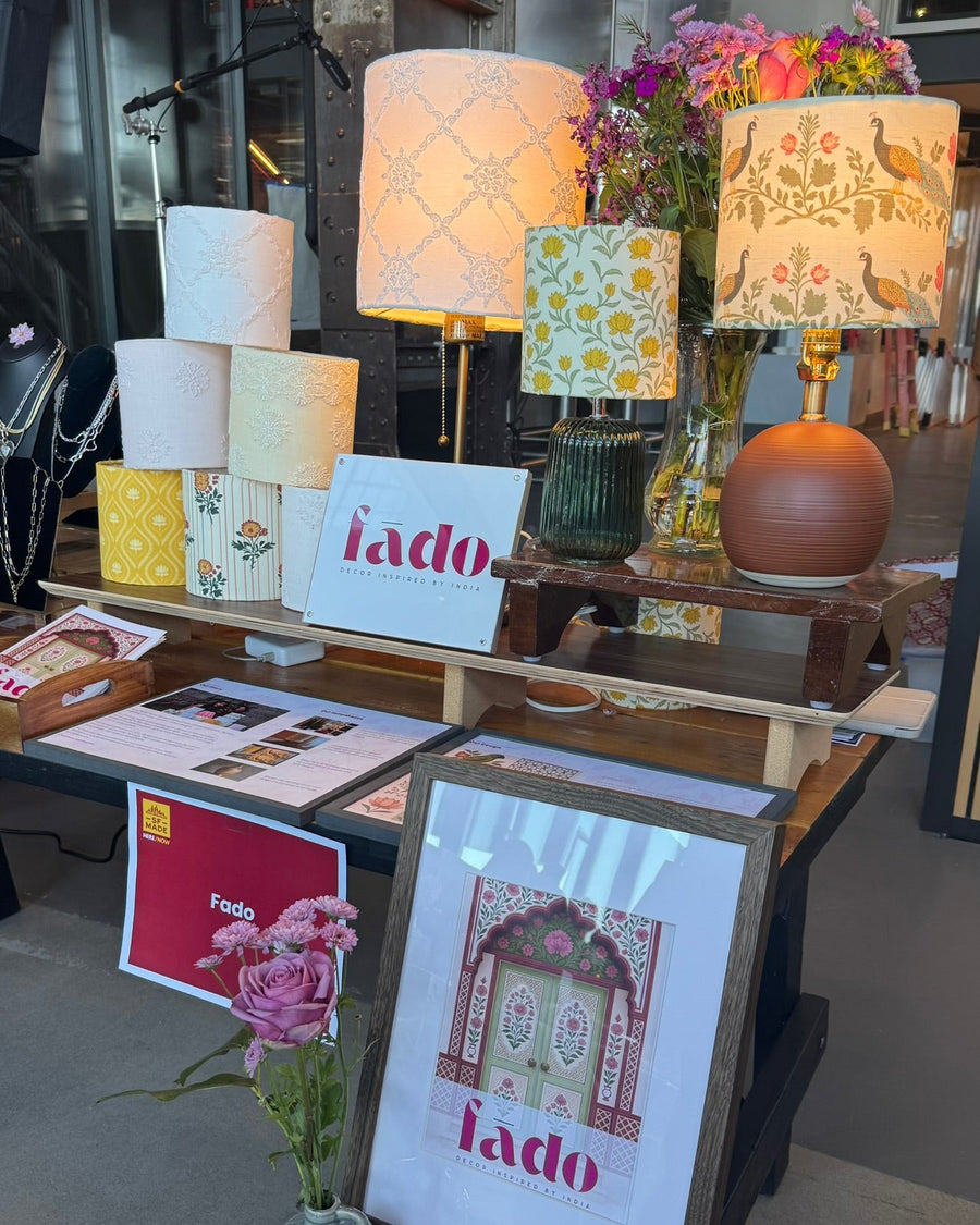 A retail display of home decor featuring multiple patterned table lamps with floral and geometric designs, a terracotta orb lamp, and framed prints with the brand name 'lado' on a wooden table in a store interior.